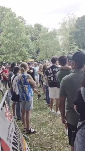 A crowd of people are gathered in a park-like setting. They are standing in a line, with some individuals interacting with each other. The environment appears to be outdoors with trees and greenery visible. A temporary fence or barrier is present in the foreground, and a sign with text is attached to it. A tent is also visible on the left side of the frame, with some equipment underneath it, possibly indicating a performance or event. The people are casually dressed, suggesting a relaxed atmosphere. The lighting indicates it is daytime. The video appears to be shot from a slightly elevated angle, looking down on the scene.