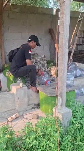 A man wearing a black t-shirt and black pants with a black backpack is sitting on a concrete ledge and using a red and white tool on a clear plastic bottle with a blue cap. He is looking down at the bottle. Another man wearing a white tank top and black and white striped pants is holding a large clear plastic bottle with a blue cap. A third man wearing a black long-sleeved shirt and khaki pants is holding a similar tool. The men are near a structure with a corrugated metal roof and concrete block walls, surrounded by green foliage and grass. There are many large clear plastic bottles filled with water stacked on the ground. The scene appears to be outdoors in daylight.