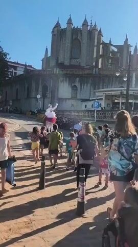 A group of people walk down a sun-drenched street towards a large, stone building with pointed spires. Many of the people appear to be children and are accompanied by adults. Some of the adults are carrying bags or pushing strollers. In the distance, a large, white inflatable figure stands tall. The ground is a mixture of dirt and pavement, with shadows cast by the people and nearby objects. A street sign with a parking symbol is visible on the building.