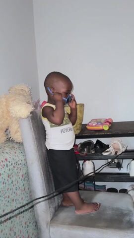 A young, barefoot toddler is putting on sunglasses in a corner of a room. The child is wearing a white tank top with a yellow trim and the words "LIMITED EDITION FUTURE HERO" on the front, and black shorts. He is standing next to a gray cushion or small mattress leaning against a white wall. A teddy bear is visible behind the cushion. To the child's right is a small, dark-colored shelf unit cluttered with shoes and a pink toy. On the shelf above, there is also a gold vase and various other items. The child adjusts the blue sunglasses on his face and then looks to his right. He raises his hand and makes a gesture as if saying something. He then turns his back to the camera.