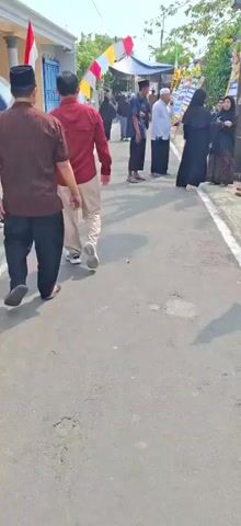 The video shows a group of people gathered outdoors on a paved street. Some people are walking away from the camera, while others are interacting in small groups. Many of the individuals are wearing traditional Islamic clothing, including hijabs and long robes. There are flags and banners visible in the background, suggesting a celebratory or ceremonial occasion. The environment appears to be a residential area with houses and trees. The lighting suggests it is daytime.