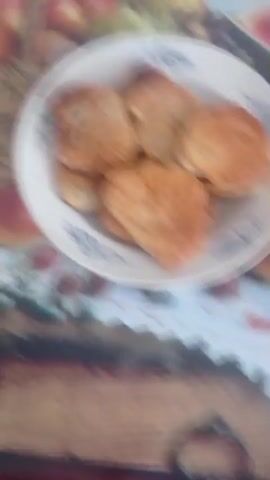 The video shows a plate of golden brown pancakes arranged on a white plate with blue decorative trim. The plate is set on a table covered with a colorful tablecloth featuring images of fruits and lace. The pancakes vary in shape and size, with some appearing slightly thicker and others flatter. The lighting suggests it might be indoors, possibly in a kitchen or dining area. The overall impression is one of a home-cooked meal.