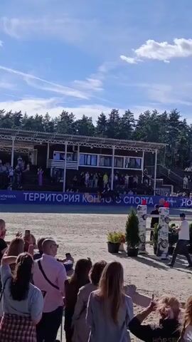 The video shows a crowd of people watching a horse jumping event. Spectators are standing and sitting in bleachers, with a rider on a horse jumping over an obstacle in the arena. A banner with the word "SPORT" is visible in the background. The weather appears to be sunny.