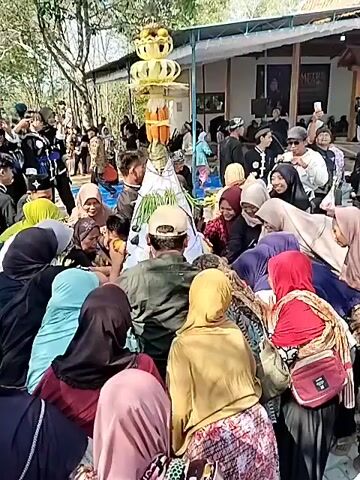 A crowd of people are gathered outdoors, with many wearing traditional clothing, specifically headscarves and loose-fitting shirts. In the center of the gathering, a cone-shaped structure adorned with what appear to be fruits or offerings is being presented. Several individuals in uniform, possibly associated with a martial arts group given their attire, are also present. The environment appears to be a village or a rural area, with wooden structures and trees visible in the background. The overall atmosphere suggests a cultural or religious ceremony.