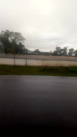 The video presents a blurry, vertical-oriented view of an outdoor scene, recorded from a moving perspective. The primary visual is a dark asphalt road, which appears damp or wet, suggesting recent rain. Along the left side of the road, a strip of green vegetation, including bushes and grass, is visible, with indistinct trees and possibly a fence or wall in the distant background. The sky is overcast and bright grey, suggesting a cloudy day, early morning, or late afternoon.

No identifiable people or specific objects are clearly discernible due to the extreme blurriness and lack of focus throughout the short clip. A distinct and continuous sound of coughing is heard from the beginning to the end of the video. There is no visible text. The setting appears to be a rural or semi-rural area, consistent with the provided context of Nduma, Kenya.