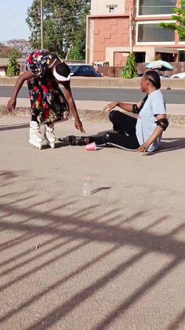 Here's a description of the video:

The video shows a sunny outdoor scene with people roller skating on a concrete surface near a road. A woman wearing a light blue shirt and black leggings with white stripes has fallen and is on her hands and knees. She's wearing roller skates and knee and elbow pads. Another woman in a colorful floral dress and roller skates is crouched down, appearing to help or check on the fallen woman. A third person, also wearing roller skates, approaches the scene. There are small, clear plastic cups scattered on the ground near the fallen woman. In the background, there's a road with cars, a building with a distinct tiered facade, and some trees. The shadows of trees are cast across the pavement.