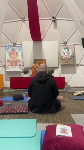 The video depicts a man sitting in a cross-legged posture on a meditation cushion inside a white geodesic dome-shaped structure. He is wearing a dark jacket and shorts, facing a table adorned with colorful cloths, flowers, and religious images, possibly Buddhist or Hindu. Two large posters featuring religious figures hang on the walls behind the table. In the foreground, there are several yoga mats and meditation cushions of different colors. A woman walks across the room, carrying a bag, passing behind the seated man. The room appears to be set up for a meditation or yoga session. A red banner hangs from the ceiling, and a book lies on a cushion in the foreground.