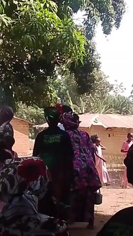 The video shows a group of people gathered in what appears to be a rural setting, possibly a village. There are trees, and simple buildings with brick walls and metal roofs are visible in the background. Several adults are standing around, some wearing traditional clothing and headwraps. One person has a t-shirt on with writing that appears to say "KAVIN MAGNA" on the back. A young girl in a pink dress is walking nearby. The overall scene suggests a social gathering, perhaps a celebration or community event. The camera seems to be held by someone within the group, capturing a partial view of the surroundings and the people present.