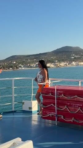A woman with dark hair stands on the deck of a ferry, looking out at a coastal town. She is wearing a floral-print top and orange shorts. The ferry has red life preservers stacked on the right side of the frame. The water is blue, and the sky is clear.