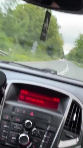 The video shows a first-person view from inside a car driving on a narrow, paved road. The car's dashboard is visible, with the radio displaying "AUX Input Device" and a temperature of 14°C. An "AIR SPICE" air freshener hangs from the rearview mirror. The road is surrounded by green trees on both sides, and the sky is overcast. Another car is visible in the distance, traveling in the same direction. The driver is not visible, but presumably behind the wheel, operating the car. Music is playing loudly in the background.