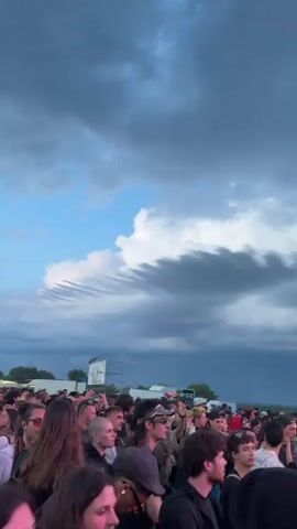 The video shows a music festival, with a crowd of people gathered in front of a large sound system. The speakers are stacked high and decorated with graffiti. There are flashing lights and the sky is cloudy. The music is loud and the crowd is lively. The video appears to be taken from the perspective of someone in the crowd.