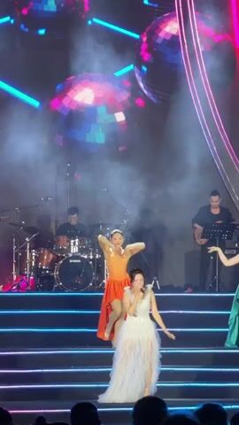 This video captures a vibrant night concert performance on an elaborate outdoor stage. The central focus is a female singer, dressed in an elegant white gown, holding a microphone and performing in front of a live audience. She is flanked by two female dancers, one in an orange dress and another in a green dress, both actively performing choreography.

Behind the performers, a live band is visible, consisting of a drummer, a guitarist, and another musician possibly on keyboard or percussion. The stage itself is multi-layered with steps, all illuminated by bright pink and blue neon lights. Large LED screens in the background display dynamic, colorful patterns and several shimmering disco balls, adding to the festive atmosphere. More physical disco balls are also suspended above the stage. Powerful spotlights beam down from above, cutting through atmospheric haze or smoke on the stage. Metal scaffolding structures are visible on either side of the main stage setup.

The text "MĂNG ĐEN KHÁT VỌNG VƯƠN XA" is prominently displayed on the large LED screen, indicating the event's location as Măng Đen and suggesting a theme of ambition or reaching out ("khát vọng vươn xa"). The audience, mostly visible as silhouettes in the foreground, watches the performance intently. The overall scene is energetic and visually rich with dynamic lighting and stage design.