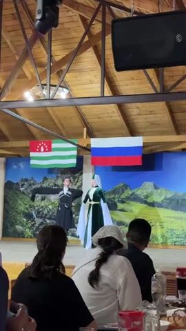 The video shows a stage performance with two dancers in traditional attire. One dancer wears a green outfit with a white headdress, and the other wears a black coat with a black hat. They are performing in front of a backdrop depicting a mountainous landscape. Two flags, one green and white with red stars and the other the Russian flag, hang behind the dancers. The audience, consisting of several people, is seated and facing the stage. Some audience members are clapping and looking at the performance. The stage is lit by spotlights. The environment appears to be an indoor venue with a wooden ceiling and exposed beams.