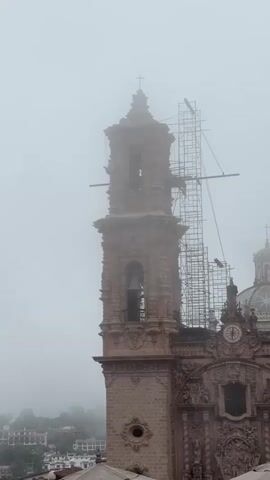 A close-up vertical shot shows the upper portion of a stone bell tower partially obscured by fog. The tower is ornate, with intricate carvings and a bell visible in an archway. Scaffolding surrounds the upper section of the tower, and a few figures can be seen on it. To the right of the tower, a portion of a dome and the facade of a building, also adorned with decorative elements and a clock, are visible. In the background, the fog creates a hazy view of distant buildings and hills.