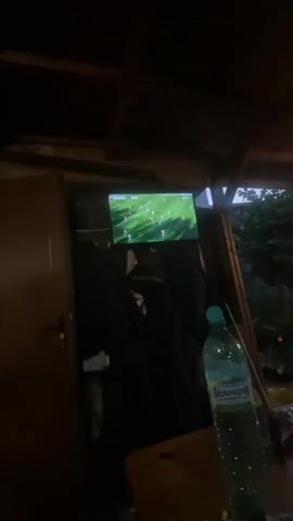 A low-light, vertical-format video shows a television screen displaying a soccer match, positioned in a dim, possibly outdoor or semi-outdoor setting. Dark clothes hang below the screen. To the right, a clear plastic water bottle with a blue cap and a white label with Cyrillic text sits on a wooden surface. The background beyond the structure shows dark foliage and a dark sky.