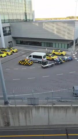 The video shows an aerial view of an airport tarmac. Several yellow taxis are parked on the left side of the screen, next to a building with large glass windows. In the center of the screen, a white van is parked, and next to it, a small tow truck is pulling several carts loaded with luggage. Another yellow taxi with a black and white checkered pattern is parked near the van. The ground is asphalt, marked with white lines. A concrete barrier with a metal fence runs along the bottom of the screen. The building in the background appears to be a terminal, with the reflection of the sky and clouds visible on its windows. The lighting suggests it is daytime, possibly late afternoon given the shadows.