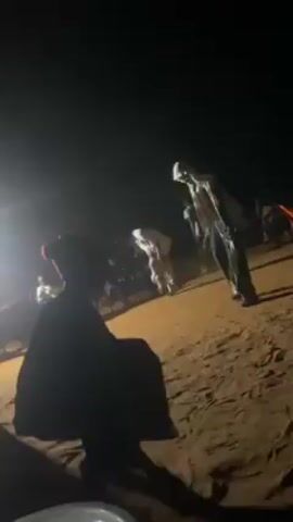 The video shows an outdoor gathering at night, likely a celebration.  A person wearing a red hat and a dark outfit is crouching in the foreground, partially obscuring the view. In the middle ground, several people are dancing on a sandy or dusty surface, illuminated by dim light sources. Some dancers wear white or light-colored clothing, which stands out against the darkness. The background is dark and blurry, suggesting the presence of more people and possibly some structures or vehicles, though they are not clearly visible.  The general atmosphere suggests a festive, though dimly lit, social event.