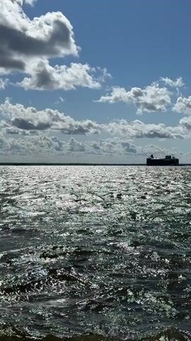 The video shows a large ship sailing on a body of water. The sun is shining, and there are clouds in the sky. The water is choppy, and the sun is reflecting off the waves. The ship is far in the distance.