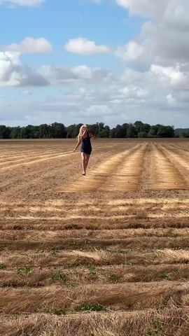 A young woman with blonde hair runs through a field of harvested crops. She wears a dark blue dress and is barefoot. The field is dry and golden, with rows of harvested plants stretching into the distance. A line of trees forms the horizon, and the sky is blue with scattered white clouds. The woman runs forward, then spins around with her arms outstretched.