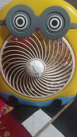 The video shows a close-up, static shot of a yellow fan designed to look like a Minion character from the Despicable Me franchise. The fan has large, gray goggle-like features with white and black circular eyes above the fan grill. The fan blades are white and are arranged in a spiral pattern. In the center of the fan blades is a white knob with the brand name "SANEX" printed on it. The fan has a blue base with two black feet. The background is blurred but appears to include a red patterned cloth on the left and a checkered floor on the right. There are no people or actions depicted in the video.