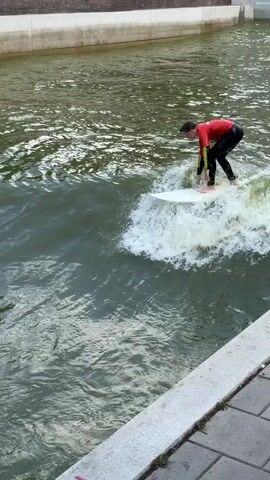 A person is surfing on a white surfboard in a river. The surfer is wearing a red long-sleeved rash guard with black sleeves and black full-length leggings. They are positioned on the board, facing away from the camera, riding a small, white-capped wave. The river water appears murky and green. The background shows a concrete embankment with some graffiti visible on the lower portion. The embankment is topped with a lighter colored concrete structure. The overall scene captures a moment of river surfing.