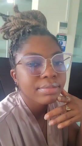 The video shows a close-up, head-and-shoulders view of a Black woman wearing large, cat-eye glasses with a light purple tint. Her hair is styled in dreadlocks gathered into a bun on top of her head. She is wearing a light pinkish-mauve satin top and several rings on her fingers, including one on her right index finger and another on her right ring finger. She also has geometric earrings. The woman is looking down and to her right in some shots, and directly at the camera in others, with her hand resting on her chin. The background appears to be an indoor office environment with a reflective glass partition, a desk, and a sign that says "Save Energy Switch Off".
