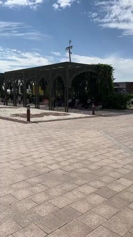 A person is walking along a paved walkway next to a row of connected, open-sided pergolas with ornate metalwork. Some people are sitting on benches under the pergolas, which are partially covered in greenery. The ground beneath the pergolas is covered in small white stones. The sky is blue with scattered clouds, and the sun is shining. There are lampposts and some metal fencing visible.