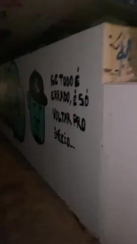 The video pans across a graffiti-covered wall in a dimly lit, enclosed space that appears to be a tunnel or underpass. The wall is white and serves as a canvas for various spray-painted artworks. On the right side of the wall, black spray paint forms lettering that reads "SE TUDO É ERRADO, É SÓ VOLTAR PRO INÍCIO...". To the left of this text, a cartoonish, teal-colored face is depicted wearing a black baseball cap with a white Yankees logo. The face has a neutral expression, with simple lines for eyes, nose, and mouth, and a goatee. Further to the left, more graffiti is visible, including large, bubbly letters in teal with red accents, and other illegible tags. The lighting is dim and uneven, casting shadows and highlighting the textured surface of the wall. The floor of the space appears to be concrete.