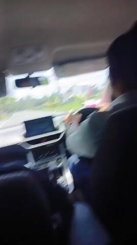 The video shows a view from inside a car driving on a paved road. The driver, wearing a white shirt and blue pants, is visible along with at least one passenger in the back. The dashboard features a digital display and climate control buttons. Through the windshield, the road ahead shows light traffic including a motorbike and a van, as well as greenery on either side. The car has sun visors still covered in protective plastic.