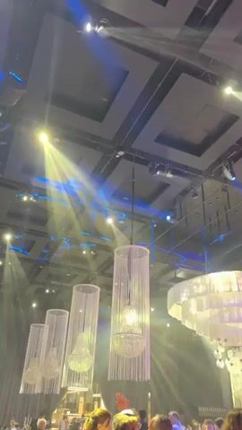 The video displays an upward-angled view of a large event space with a high ceiling, filled with dynamic lighting effects. Multiple spotlights cast sweeping beams of yellow, blue, and red light across the room, with haze in the air making the light rays visible. Several decorative chandeliers hang from the ceiling. Four tall, cylindrical chandeliers with internal lights are visible on the left, changing color with the room's lighting. A large, tiered, circular chandelier with multiple internal lights is prominent on the right, also changing color. Speakers are suspended from the ceiling grid. Out-of-focus people are visible in the lower portion of the frame, suggesting an audience or guests at an event. A camera on a tripod is briefly visible in the lower right foreground. Upbeat music is audible throughout the video.