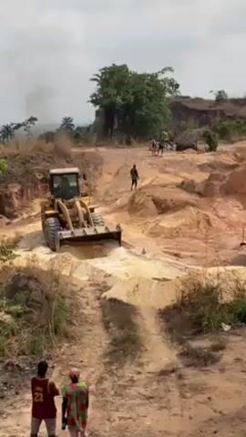 The video shows a yellow bulldozer traversing a sandy, uneven terrain, presumably at a quarry or mining site. Two people stand in the foreground, observing the machinery. In the distance, more individuals are visible near a rudimentary shelter. The area is characterized by sparse vegetation, exposed earth, and evidence of excavation. There's some smoke rising in the background, possibly from a distant fire. The overall setting appears to be a rural, underdeveloped area, potentially in a tropical or subtropical region.