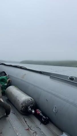The video shows the interior of a gray inflatable boat moving through calm water under a cloudy sky. In the boat, there are diving tanks, a net, and plastic bags. The water is dark and reflective, and the sky is a pale gray. In the distance, there is a wooded shoreline.