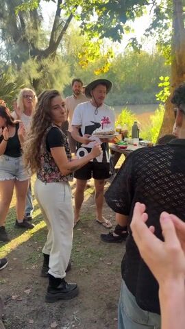 A group of approximately a dozen people gather in an outdoor setting that appears to be a park or garden area. A person wearing a bucket hat holds a cake with a lit candle, and the group is clapping and singing "Happy Birthday." The birthday celebrant dances and smiles. Some of the people appear to be sitting at a table. The environment includes trees, greenery, and a river in the background. The atmosphere suggests a casual and joyous celebration.
