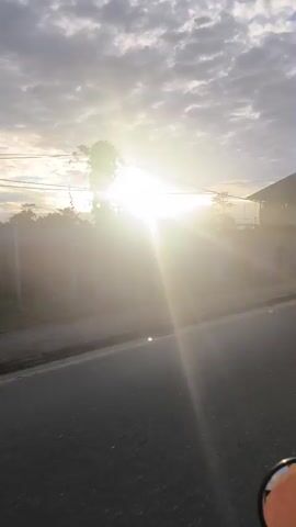A dash cam captures a roadside view with the sun shining brightly. The camera pans across a road, a sidewalk, a fence, and a building. The sun's glare obscures much of the scene, but glimpses of trees and power lines are visible. The building appears to be under construction or in disrepair, with scaffolding visible on its upper floors. The lower level has storefronts with signs and graffiti. The video ends with another view of the sunlit roadside.