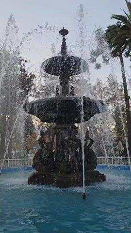 A multi-tiered ornate fountain with water spraying from the top and sides is the central focus. The fountain is made of dark material, possibly bronze or stone, and features sculpted figures around its base. It sits in a bright blue pool of water, surrounded by lush green trees and a tall palm tree. The sky above is overcast, suggesting a daytime scene. The video is filmed at eye-level, capturing the dynamic movement of the water and the intricate details of the fountain. There are no people visible in the frame.