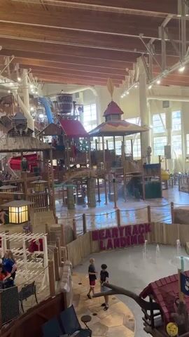 The video shows an indoor water park with a wooden roof structure and large windows. In the foreground, there is a shallow water play area with a wooden fence and the words "HEARTTRACK LANDING" displayed in large pink letters on the fence. Several children are present in this area. To the right, a larger swimming pool is visible with many people, including children and adults, swimming and playing in the water. In the background, there are water slides and other water park attractions. Tables and chairs are set up around the pool area, and there are also some buildings that appear to be part of the water park facilities. The overall atmosphere seems to be one of a lively and fun recreational environment.