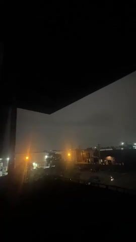 The video shows a nighttime view of an industrial area with buildings and lights. The sky is dark and overcast, suggesting potential bad weather, and there is a distant flash of lightning. The foreground is dark, with what appears to be a railing or balcony structure.