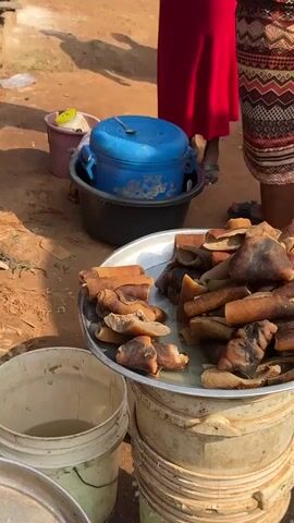 The video captures a bustling outdoor market scene, likely in a warm climate due to the sandy ground and bright sunlight. In the foreground, a large metal platter is filled with cooked animal parts, appearing to be pig trotters or similar cuts, with visible skin and bone. These are arranged in piles, suggesting they are for sale. To the left and right of the platter are large white plastic buckets; one on the left contains a murky yellow liquid. Behind the platter, a person wearing a red dress and a patterned garment stands near a blue lidded pot placed inside a larger black container. Another person, partially visible on the right, also wears a patterned garment. A third person, wearing a red and white patterned top and a red skirt, is seen bending down and reaching into a pink bucket. The ground is dirt and sand, with scattered debris and a rough stone structure on the far left. The overall impression is one of an active food vendor or market stall.