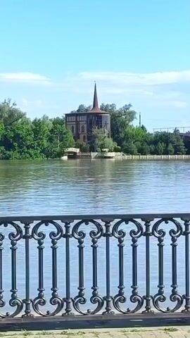 A picturesque view of a serene lake unfolds, with a quaint building featuring a towering spire nestled amidst lush trees on the opposite shore. A red boat glides gracefully across the water, adding a touch of dynamism to the tranquil scene. In the foreground, a beautifully crafted metal fence with intricate scrollwork stands, framing the captivating landscape. The sky above is a clear, azure blue, dotted with a few wispy clouds, completing this idyllic image.