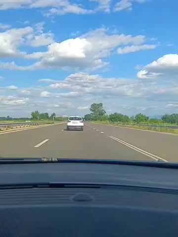 The video shows a view from inside a car driving on a highway. The perspective is from the dashboard, looking through the windshield. Ahead on the highway, there is a white car traveling in the same direction. To the left of the white car, a dark-colored car is visible. The highway has clear lane markings and is bordered by a guardrail on the left and trees and vegetation on the right. The sky above is blue with scattered white clouds. The road appears to be in good condition, and the lighting suggests it is daytime.