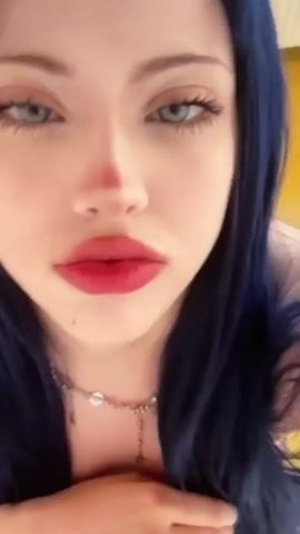 A close-up video shows a person with blue hair and striking blue eyes looking directly at the camera. They have bold red lipstick on and are wearing a necklace with a pendant. The person's left hand is visible, resting on their chest. The background is blurred, with hints of yellow suggesting an indoor setting. The video is short and appears to be a selfie or a short recorded message.