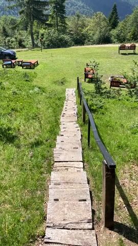The video shows a pair of outdoor wooden stairs descending from a grassy area with picnic tables toward a mountainous landscape. On the left, a dark-colored car is parked near some of the picnic tables. Several red and blue picnic tables with benches are scattered across the green lawn. In the background, lush green trees and rolling mountains under a bright blue sky are visible. A black metal railing lines the right side of the stairs. The camera angle is looking down the stairs, giving a sense of depth and perspective.