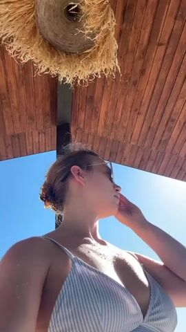 This video features a close-up, low-angle shot of a young woman wearing a white and light blue striped bikini top and sunglasses. She is looking off to the right, with her hand raised near her face, shielding her eyes from the sun. Her hair is pulled back, with some strands loose around her face. Water droplets are visible on her skin, suggesting she may have recently been swimming.

She appears to be seated beneath a thatched roof structure, which casts a shadow and provides some shade. The background consists of a clear, bright blue sky, indicating a sunny day. The wooden planks of the roof are visible above her, and a circular, fringe-like object is also hanging from the roof.

The overall mood is relaxed and summery, possibly indicating a day at the beach or a poolside. The strong sunlight suggests it is midday or early afternoon. The location context of Rezekne, Latvia, places the scene in a specific geographical setting. There is no visible text in the video.