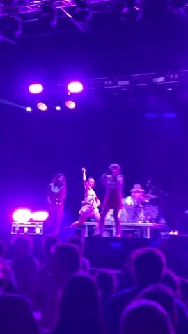 A stage performance with multiple female musicians. One woman is playing a trumpet, another is singing into a microphone, and a third is playing a guitar. A drummer is visible in the background. The stage is lit with purple and pink lights. The audience is visible in the foreground, with their backs to the camera.