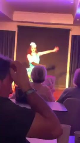 A performer in a shiny turquoise dress and a white headpiece is dancing on a small stage in front of an audience. The lighting changes colors from purple to green to blue. The audience is seated at tables and consists primarily of people with short grey hair. The video is shaky and appears to be taken from a seat in the audience.