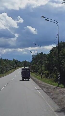 A dark-colored truck with "OZON" written on its side drives down a two-lane asphalt road. The road is bordered by trees on both sides, with power lines and streetlights visible along the right side. The sky is partly cloudy, with patches of blue and gray. The camera appears to be mounted on a vehicle following the truck.