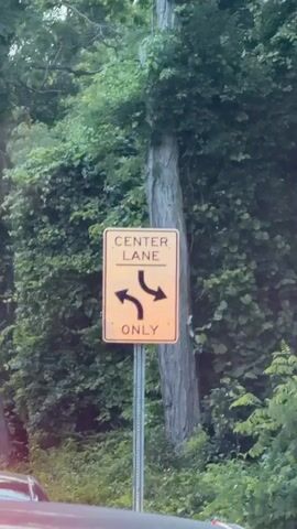 A close-up, shaky video shows a traffic sign. The sign is orange with black text and arrows. It reads "CENTER LANE ONLY" and has two curved arrows pointing left and right, indicating that vehicles in the center lane can turn in either direction. The sign is mounted on a metal pole in front of a lush green background of trees and foliage. A portion of a car is visible in the lower left corner of the frame, out of focus. The video appears to be filmed from a moving vehicle.