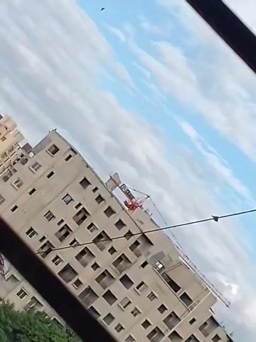 The video shows a tilted view of a tall apartment building under construction. The building has many windows that are dark, suggesting they are either empty or unfinished. A large red crane is visible at the top of the building, indicating ongoing construction work. In the background, the sky is visible with scattered clouds. Part of a dark, horizontal object, possibly a wire or railing, runs across the foreground, obscuring parts of the building and trees. Green trees are visible at the bottom left of the frame. The overall scene suggests an urban environment with active construction.