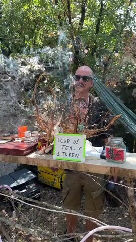 A bald man with sunglasses stands behind a wooden table outdoors, in front of a dense green forest. He has a black shirt on and khaki shorts. He is smoking, with smoke obscuring his face and rising into the air around a decorative arrangement of twigs and peacock feathers. On the table are two small white cups, one labeled "MIX," an orange plastic cup, a jar of what appears to be jam, and a sign that reads "1 CUP OF TEA - 1 TOKEN." A green hammock is visible in the background. The scene is illuminated by natural sunlight.