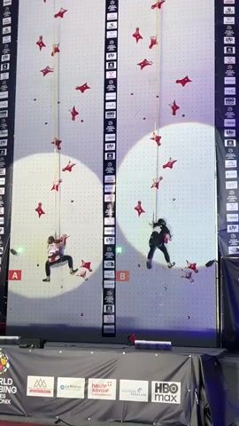 Two athletes are climbing vertically up two parallel, white climbing walls. The walls are covered with red climbing holds. Each athlete is attached to a rope that hangs from the top of the wall. Both athletes are wearing climbing gear, including harnesses. The wall on the left has a white circle projected onto it, highlighting the athlete on that wall. The wall on the right also has a white circle projected onto it, highlighting the athlete on that wall. The area below the climbing walls is a stage with banners and logos. The background appears to be outdoors, with mountains visible in the distance. The lighting suggests it is evening or nighttime. The scene is likely a competitive climbing event.
